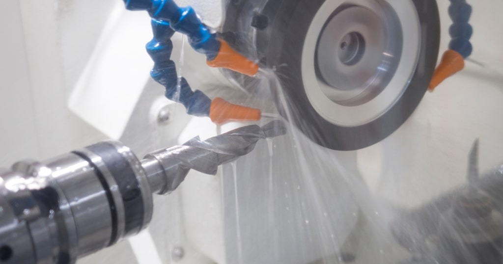Närbild av en spiralborr som slipas mot en roterande slipskiva i en CNC-slipmaskin, medan kylvätska sprutas från blå och orange munstycken.